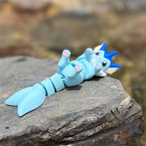 May include: A light blue articulated cat-like figurine with a fin tail, lying on its back. The figurine has pink paw pads and blue and white accents. The toy is resting on a gray rock, with a blurred natural background.