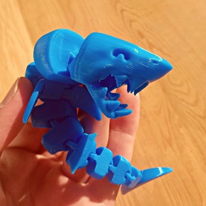 May include: A bright blue 3D printed articulated shark toy. The shark has a segmented body and a large head with a toothy grin.