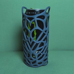 May include: A gray, open-weave can cooler holds a black and green Monster energy drink can. The cooler has a unique, abstract design with two handles at the top. The background is a solid green.