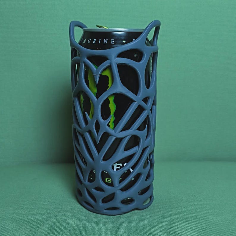 May include: A gray, open-weave can cooler holds a black and green Monster energy drink can. The cooler has a unique, abstract design with two handles at the top. The background is a solid green.