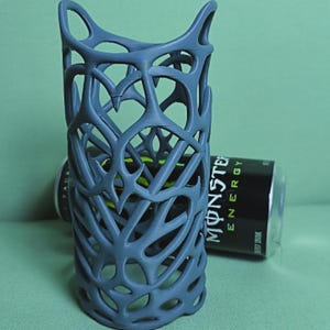 May include: A gray, 3D-printed beverage can holder with an intricate, openwork design. The holder is cylindrical and features a complex pattern of interconnected shapes. A Monster Energy drink can is visible inside the holder and another one is behind it.