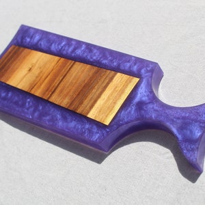 May include: A purple and wood serving board with a unique handle. The board is made of resin and has a wood inlay.