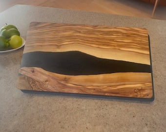 May include: A rectangular wooden cutting board with a dark, river-like epoxy resin inlay. The board features a natural wood grain pattern with light and dark brown tones. The edges are dark gray. The board is approximately 16 inches long and 12 inches wide.
