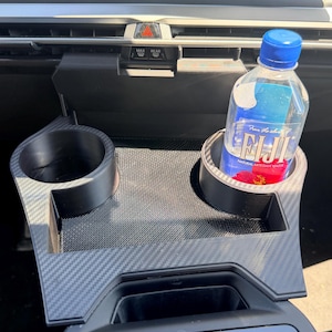 以下が含まれることがあります： カーボンファイバー柄の黒いプラスチック製カップホルダーインサート。2つの円形の開口部があり、そのうちの1つには青いキャップが付いた透明なプラスチック製のフィジーウォーターボトルが入っており、車両のセンターコンソールに配置されています。
