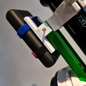 Pu&ograve; includere: Un supporto per telefono bianco stampato in 3D attaccato a un supporto per telescopio verde e bianco. Il supporto per telefono ha un power bank nero fissato con cinturini in velcro neri. Il power bank ha un cinturino in velcro blu sull'altro lato.