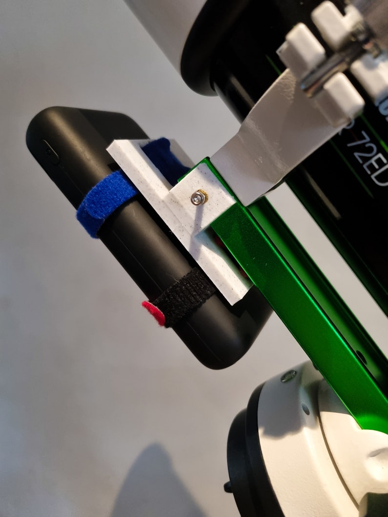 Pu&ograve; includere: Un supporto per telefono bianco stampato in 3D attaccato a un supporto per telescopio verde e bianco. Il supporto per telefono ha un power bank nero fissato con cinturini in velcro neri. Il power bank ha un cinturino in velcro blu sull'altro lato.
