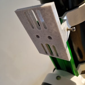 Pu&ograve; includere: Una staffa bianca stampata in 3D con tre fessure e due fori, fissata a un supporto per telescopio verde e bianco.