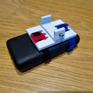 Pu&ograve; includere: Un supporto per telefono bianco stampato in 3D con un cinturino rosso e blu attaccato a una custodia per telefono nera. Il supporto ha un piccolo foro per una vite.