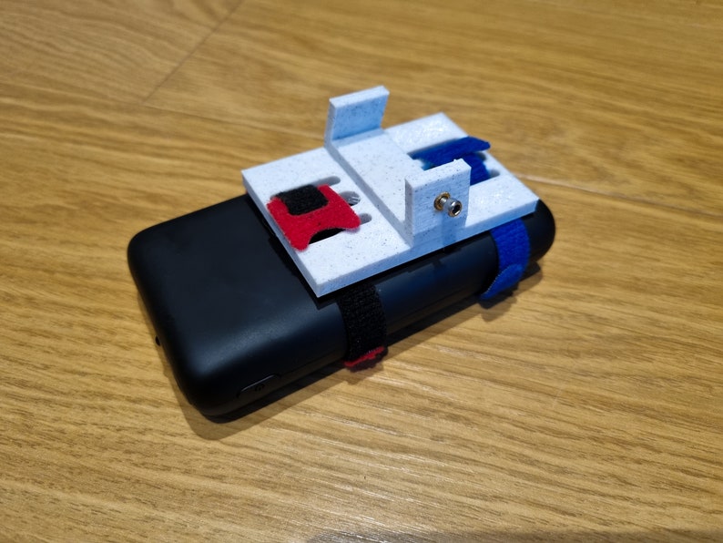 Pu&ograve; includere: Un supporto per telefono bianco stampato in 3D con un cinturino rosso e blu attaccato a una custodia per telefono nera. Il supporto ha un piccolo foro per una vite.