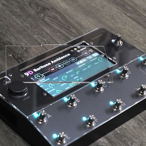 Puede incluir: Un pedal de efectos negro y plateado con m&uacute;ltiples perillas e interruptores de pie. La pantalla muestra "9D Baritone Ambience" y otras configuraciones. El dispositivo tiene luces LED azules y est&aacute; sobre una superficie de madera.