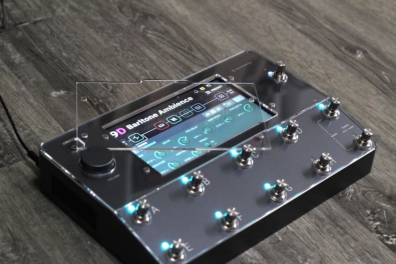 Puede incluir: Un pedal de efectos negro y plateado con m&uacute;ltiples perillas e interruptores de pie. La pantalla muestra "9D Baritone Ambience" y otras configuraciones. El dispositivo tiene luces LED azules y est&aacute; sobre una superficie de madera.