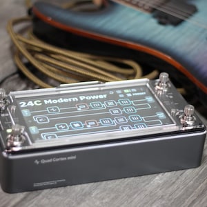 Könnte beinhalten: Ein schwarzer und silberner Quad Cortex Mini Gitarreneffektprozessor mit einem Bildschirm, der eine grafische Oberfläche anzeigt. Der Text "24C Modern Power" ist sichtbar. Eine mehrfarbige E-Gitarre und ein Kabel befinden sich im Hintergrund.