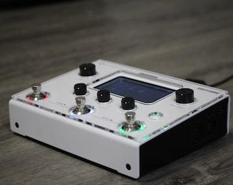 Puede incluir: Pedal de efectos de guitarra blanco con base negra y una pequeña pantalla LCD. El pedal tiene cuatro botones, dos interruptores de pedal y un cable de alimentación negro.