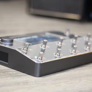 Puede incluir: Un pedal de efectos negro y plateado con m&uacute;ltiples interruptores de pie plateados y una pantalla. El dispositivo tiene forma rectangular con una parte superior transparente y una base negra. Dise&ntilde;ado para instrumentos musicales.