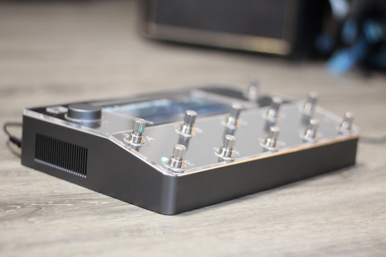 Puede incluir: Un pedal de efectos negro y plateado con m&uacute;ltiples interruptores de pie plateados y una pantalla. El dispositivo tiene forma rectangular con una parte superior transparente y una base negra. Dise&ntilde;ado para instrumentos musicales.