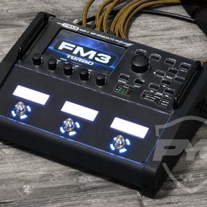 Può includere: Un processore di effetti e modellatore di amplificatore FM3 Mark II Turbo nero con display digitale centrale, tre interruttori a pedale metallici con anelli LED blu, diverse manopole di controllo, pulsanti di navigazione e cavi di ingresso.
