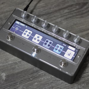 Könnte beinhalten: Ein silbergraues Effektpedal mit einem digitalen Display, das eine Effektkette anzeigt. Es hat mehrere Knöpfe und Fußschalter. Das Display zeigt den Text "868 DOOM" und eine Effektkette. Ein schwarzes Kabel ist oben eingesteckt.