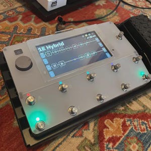 Puede incluir: Un pedal de efectos plateado con el texto "1E Hybrid" en la pantalla. Tiene m&uacute;ltiples perillas y botones plateados, con luces indicadoras verdes y azules. El dispositivo est&aacute; sobre una base negra, con una alfombra estampada en el fondo.