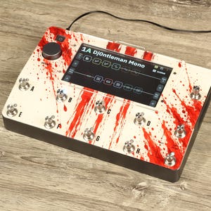 May include: A digital guitar effects processor featuring a cream-colored top panel with a red blood-splatter pattern. The device includes a central touchscreen displaying the text 1A Dj0ntleman Mono, multiple metallic footswitches labeled A through H, and a black rotary knob.
