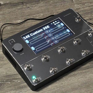 Puede incluir: Un pedal de efectos 14E Custom 3SE gris oscuro con una pantalla que muestra la configuraci&oacute;n. Tiene m&uacute;ltiples interruptores de pie plateados, una perilla de volumen e indicadores luminosos. Un cable negro est&aacute; conectado.