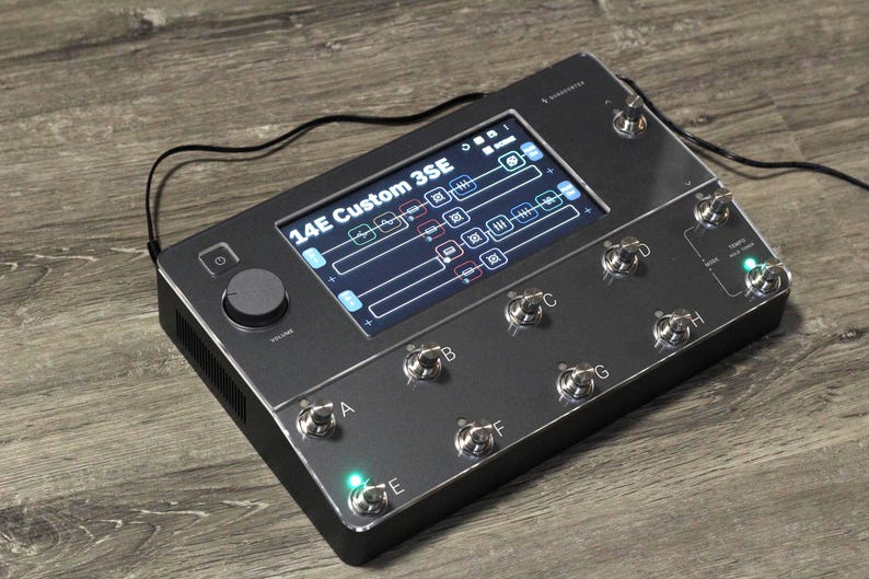 Puede incluir: Un pedal de efectos 14E Custom 3SE gris oscuro con una pantalla que muestra la configuraci&oacute;n. Tiene m&uacute;ltiples interruptores de pie plateados, una perilla de volumen e indicadores luminosos. Un cable negro est&aacute; conectado.
