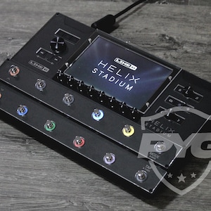 May include: A black Line 6 Helix guitar processor featuring a large central display screen with the text "LINE 6 HELIX STADIUM" visible. The unit includes multiple metallic footswitches with colored rings, a rotary knob, and various control buttons.