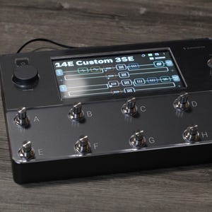Puede incluir: Un dispositivo electr&oacute;nico negro con una pantalla que muestra configuraciones, etiquetado como "14E Custom 3SE". Cuenta con m&uacute;ltiples perillas y botones plateados, con un dial central grande. El dispositivo est&aacute; sobre una superficie de madera.