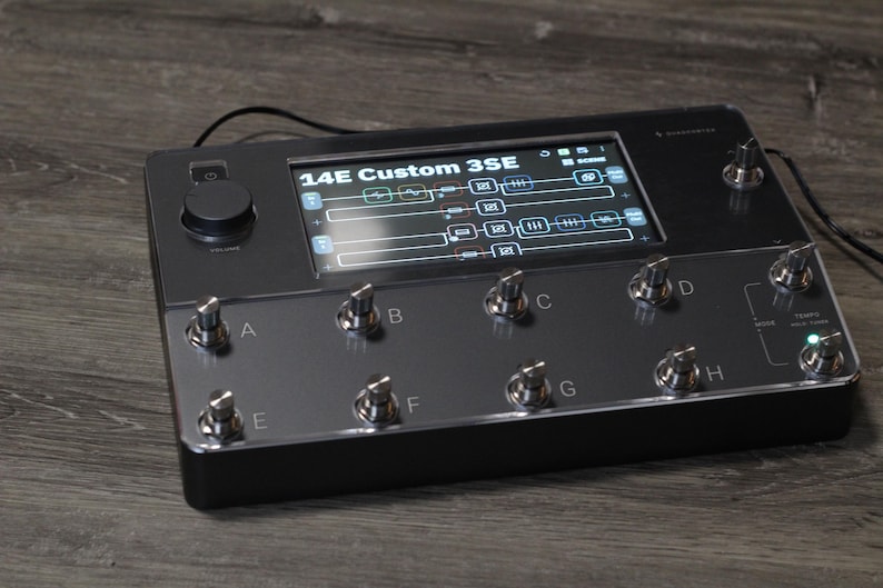 Puede incluir: Un dispositivo electr&oacute;nico negro con una pantalla que muestra configuraciones, etiquetado como "14E Custom 3SE". Cuenta con m&uacute;ltiples perillas y botones plateados, con un dial central grande. El dispositivo est&aacute; sobre una superficie de madera.