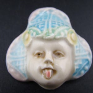 May include: A ceramic brooch featuring a sculpted face with a protruding tongue. The figure wears a blue and white patterned headpiece with yellow floral accents. The face is off-white with pinkish tones on the tongue and subtle shading around the eyes and nose, creating a three-dimensional appearance.