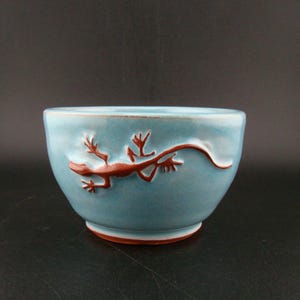 May include: A turquoise ceramic bowl with a brown lizard design. The bowl has a rounded shape and a slightly flared rim. The lizard is embossed on the side of the bowl.