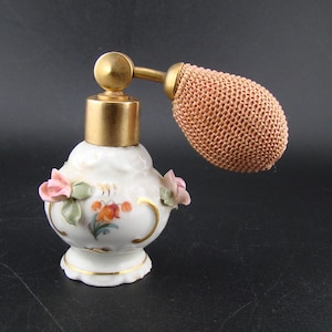 May include: A vintage white porcelain perfume atomizer with a gold-colored metal top and a mesh bulb. The atomizer is decorated with pink and green floral accents and gold trim. The bulb is a light peach color.