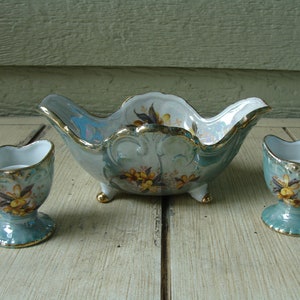 May include: A vintage porcelain serving set featuring a scalloped bowl and two egg cups. The set is painted in a pale blue-green with gold trim and yellow floral accents. The bowl has three small feet.