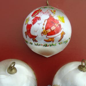 May include: Three white Christmas ornaments with a painted design of Santa Claus and a child. The ornaments have a silver loop for hanging.