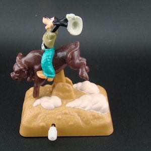 May include: A vintage toy featuring a cartoon character riding a brown bull. The character wears a green shirt, blue pants, and a cowboy hat. The toy is set on a tan base with white cloud accents.