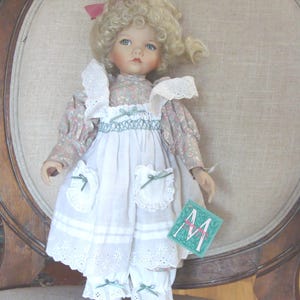 May include: A porcelain doll with blonde curly hair wearing a white dress with green and pink floral accents. The doll is holding a green tag with the letter "M" on it.
