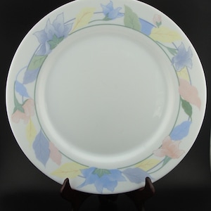 May include: White ceramic plate with a floral border featuring blue, pink, and yellow flowers and leaves. The plate is displayed on a dark brown wooden stand, showcasing its decorative design.