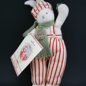 May include: A plush white bunny doll wearing a striped hat, scarf, and overalls with red stripes. The scarf reads "Joy Phil." The doll is holding a green sprig and a tag that reads "Bunnies by the Bay."