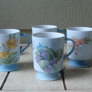 May include: Four blue and white ceramic mugs with floral designs. Each mug has a different floral pattern with yellow, blue, and pink flowers. The mugs have a blue base.