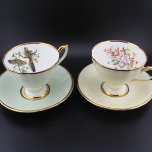 May include: Two teacups and saucers with gold trim. One saucer is light green with a bird design, the other is cream with a floral design.
