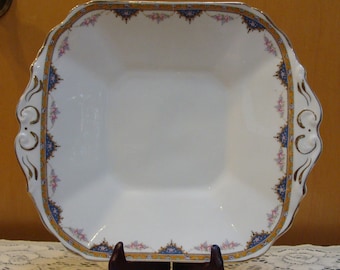 May include: A white square serving dish with a gold and blue floral border. The dish has a scalloped edge and is on a wooden stand.
