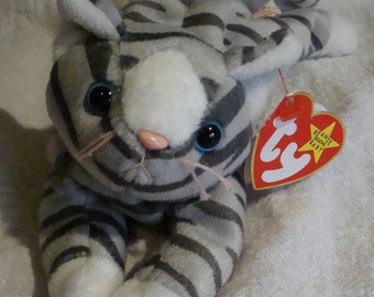 May include: A plush gray and white cat toy with black stripes. The cat has large blue eyes, a pink nose, and pink whiskers. A red heart-shaped tag with the words "Beanie Original Baby" is attached.