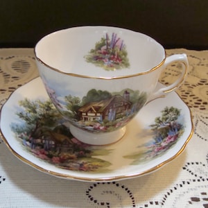 May include: A white teacup and saucer with a gold rim. The teacup and saucer feature a painted scene of a cottage in a garden with flowers and trees.