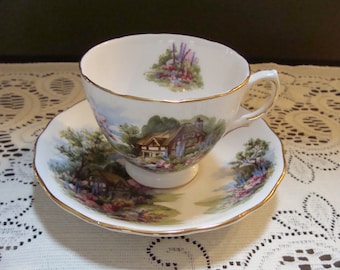 May include: A white teacup and saucer with a gold rim. The teacup and saucer feature a painted scene of a cottage in a garden with flowers and trees.