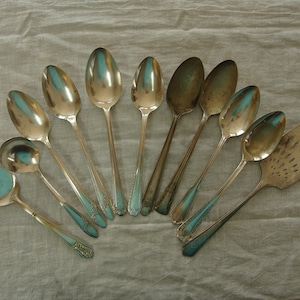 May include: A collection of ten silver spoons and a silver serving utensil with a slotted design. The spoons are arranged in a fan shape on a light beige fabric background.