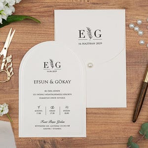 May include: Wedding stationery set featuring a white arched invitation card and envelope, both with the initials "E G" and the date "16 Haziran 2029". Gold scissors, a pen, pearls, and flowers are arranged on a wooden surface.