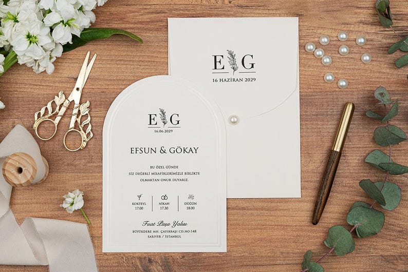 May include: Wedding stationery set featuring a white arched invitation card and envelope, both with the initials "E G" and the date "16 Haziran 2029". Gold scissors, a pen, pearls, and flowers are arranged on a wooden surface.