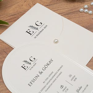 May include: Ivory wedding invitation suite with black text and a floral design. The invitation includes the initials "E G" and the date "16 HAZIRAN 2029". A pearl embellishment and gold scissors are also visible.