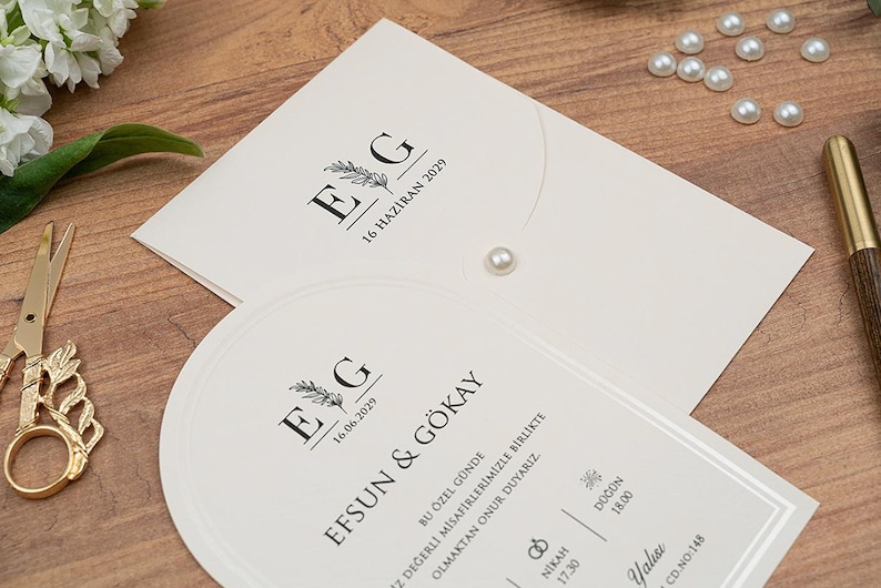 May include: Ivory wedding invitation suite with black text and a floral design. The invitation includes the initials "E G" and the date "16 HAZIRAN 2029". A pearl embellishment and gold scissors are also visible.