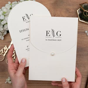 May include: Ivory wedding invitation suite with a flap closure and pearl detail. The invitation features the initials "E G" and the date "16 HAZIRAN 2029" in black. A matching fan-shaped card is visible. Gold scissors and a pen are also present.