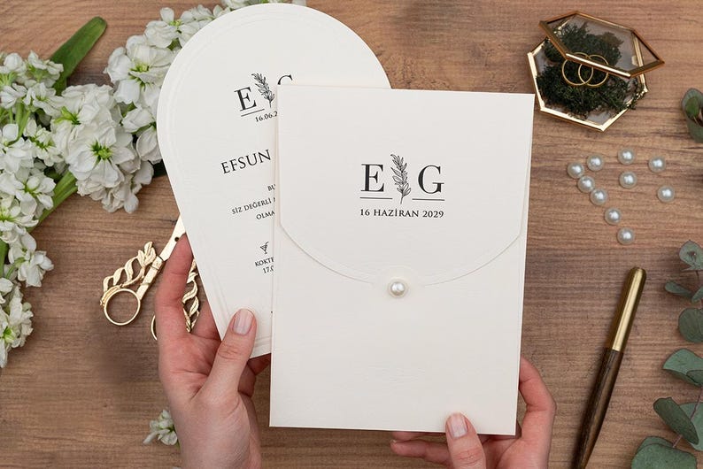 May include: Ivory wedding invitation suite with a flap closure and pearl detail. The invitation features the initials "E G" and the date "16 HAZIRAN 2029" in black. A matching fan-shaped card is visible. Gold scissors and a pen are also present.